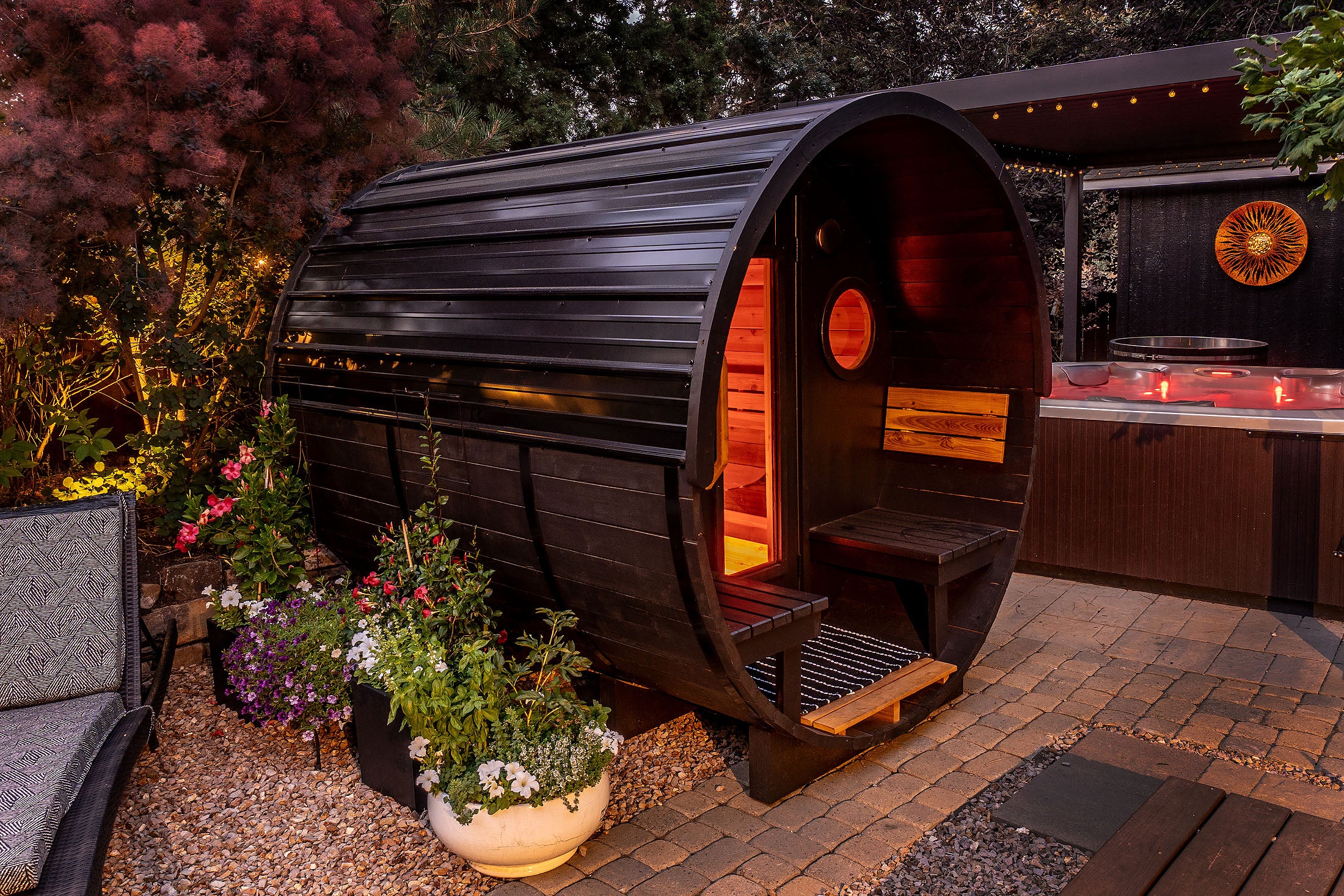 Wooden barrel-style sauna in a backyard setting with plants and a hot tub.