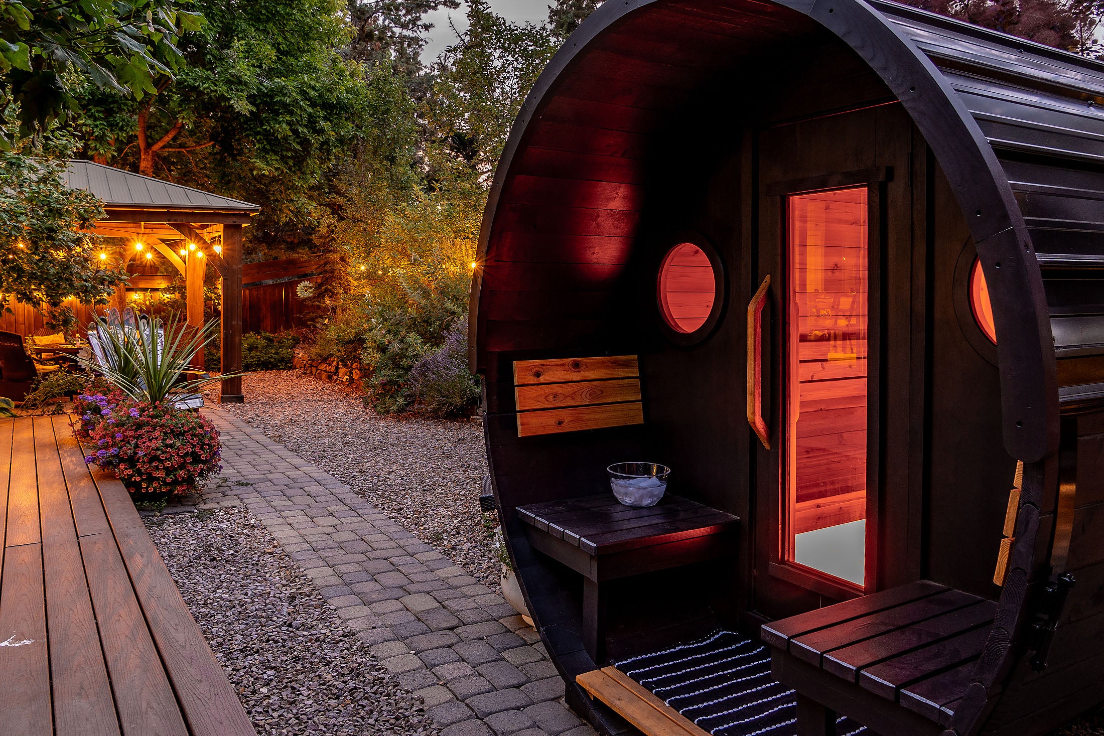 Wooden barrel-shaped sauna with dark stain in a garden setting with plants and a gazebo.