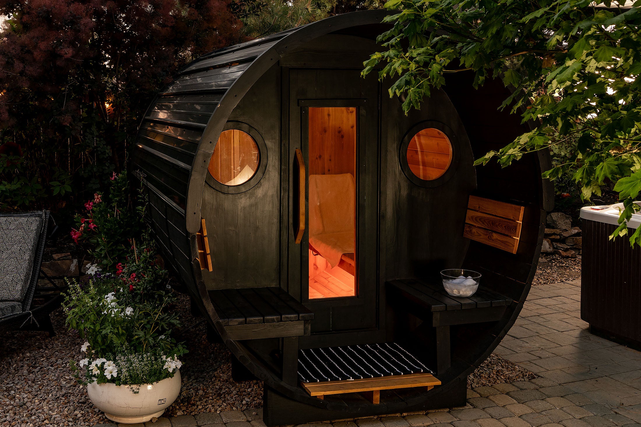 Wooden black stained cedar barrel sauna with a warm interior light, surrounded by plants and garden elements.